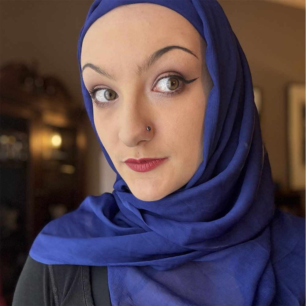A 20-something-year-old woman wearing a blue hijab looks at the camera with her head tilted. The image is black-and-white until it's hovered over. She has big green-ish eyes, and one nostril is pierced with a black stud. She is wearing dramatic makeup with a red lip, sharp eyeliner and thin brows. The background is out of focus but warm with ambient brown and orange tones.